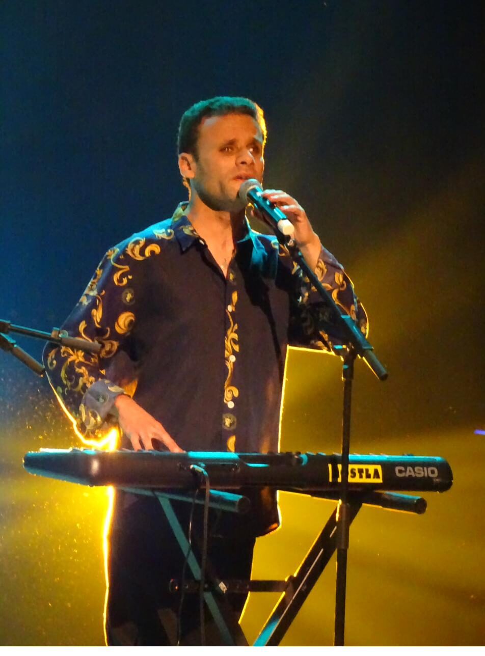 Chanteur au clavier sur scène, chemise à motifs, dans des lumières bleue et jaune
