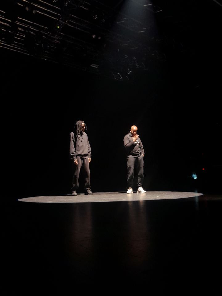 Deux interprètes debout sur un plateau sombre, dans un rond de lumière