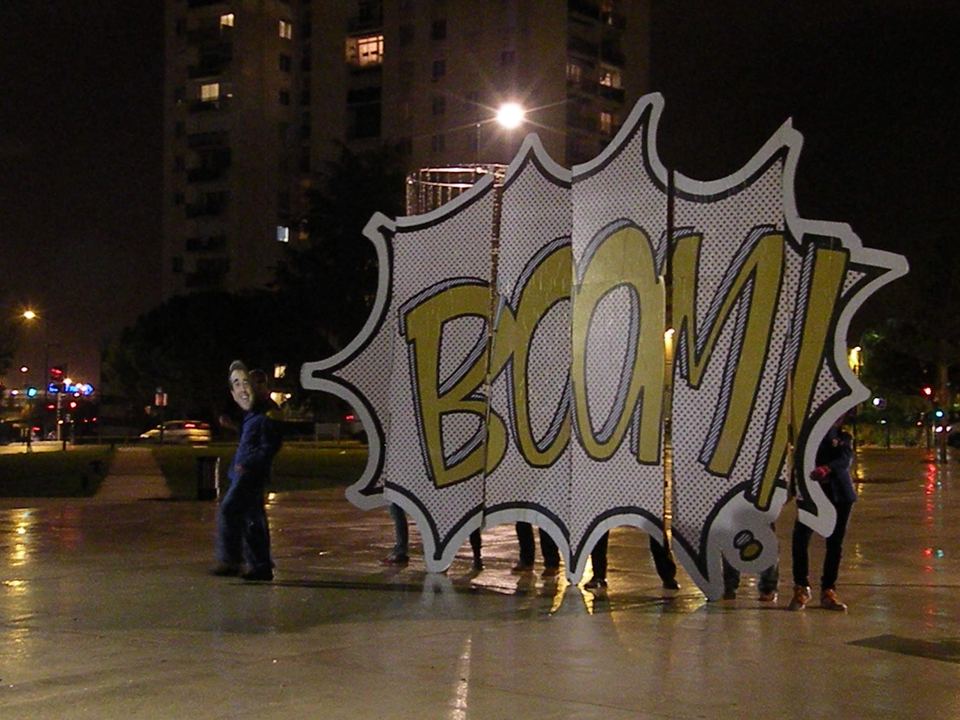 De nuit, sur une place, une grande bulle de bande dessinée « BOOM ! » et une personne masquée