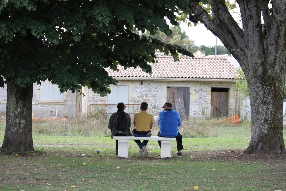 Trois personnes assises sur un banc face à une maison de pierre, sous les arbres