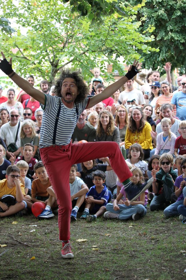 Artiste en marinière et pantalon rouge, bras écartés et jambe levée, devant un public assis sous les arbres